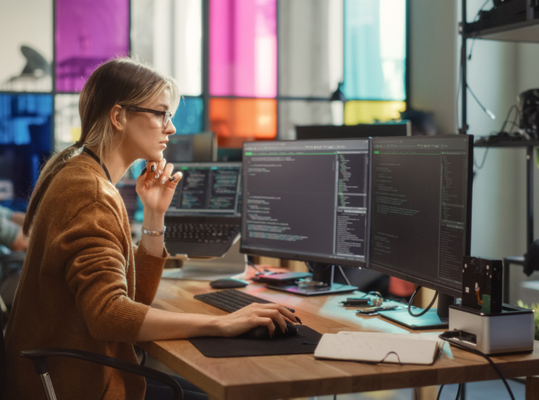 Caucasian Woman Writes Lines of Code on Desktop PC with Multiple Displays and a Laptop in Stylish Office. Professional Female Data Scientist Using Software To Analyze Information from Internet. Woman coding on a computer, representing Ceiba Law’s support for software developers and tech startups